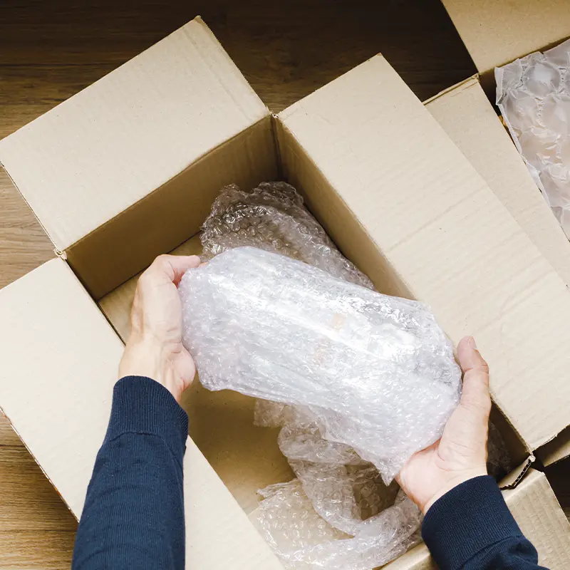 caixa-de-papelao-aberta-maos-segurando-objeto-envolto-em-plastico-bolha-visto-de-cima